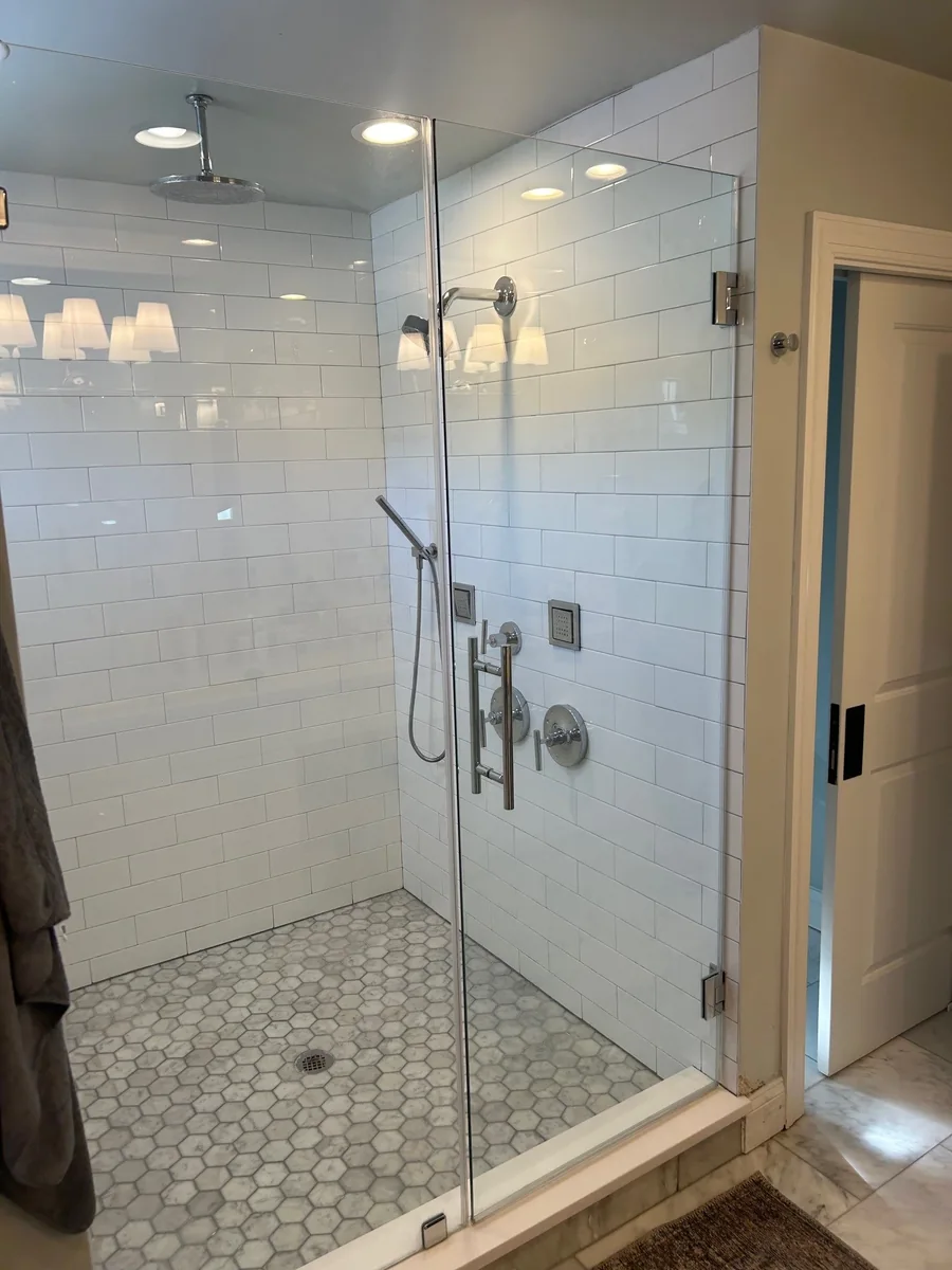 Frameless shower door and fixed glass panel reveal white subway tile and a hexagonal tile floor.