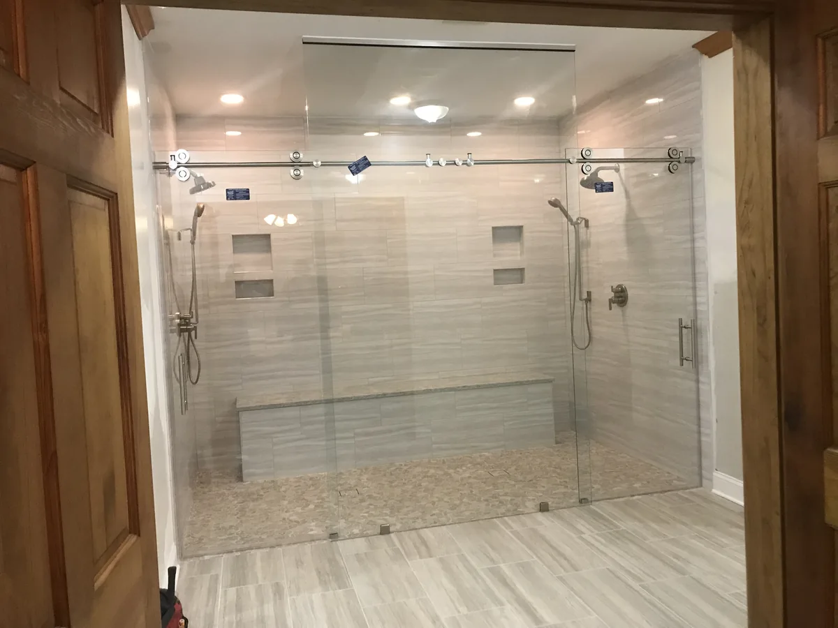 Wide sliding glass shower enclosure with exposed rollers, two shower heads, and a tiled bench.