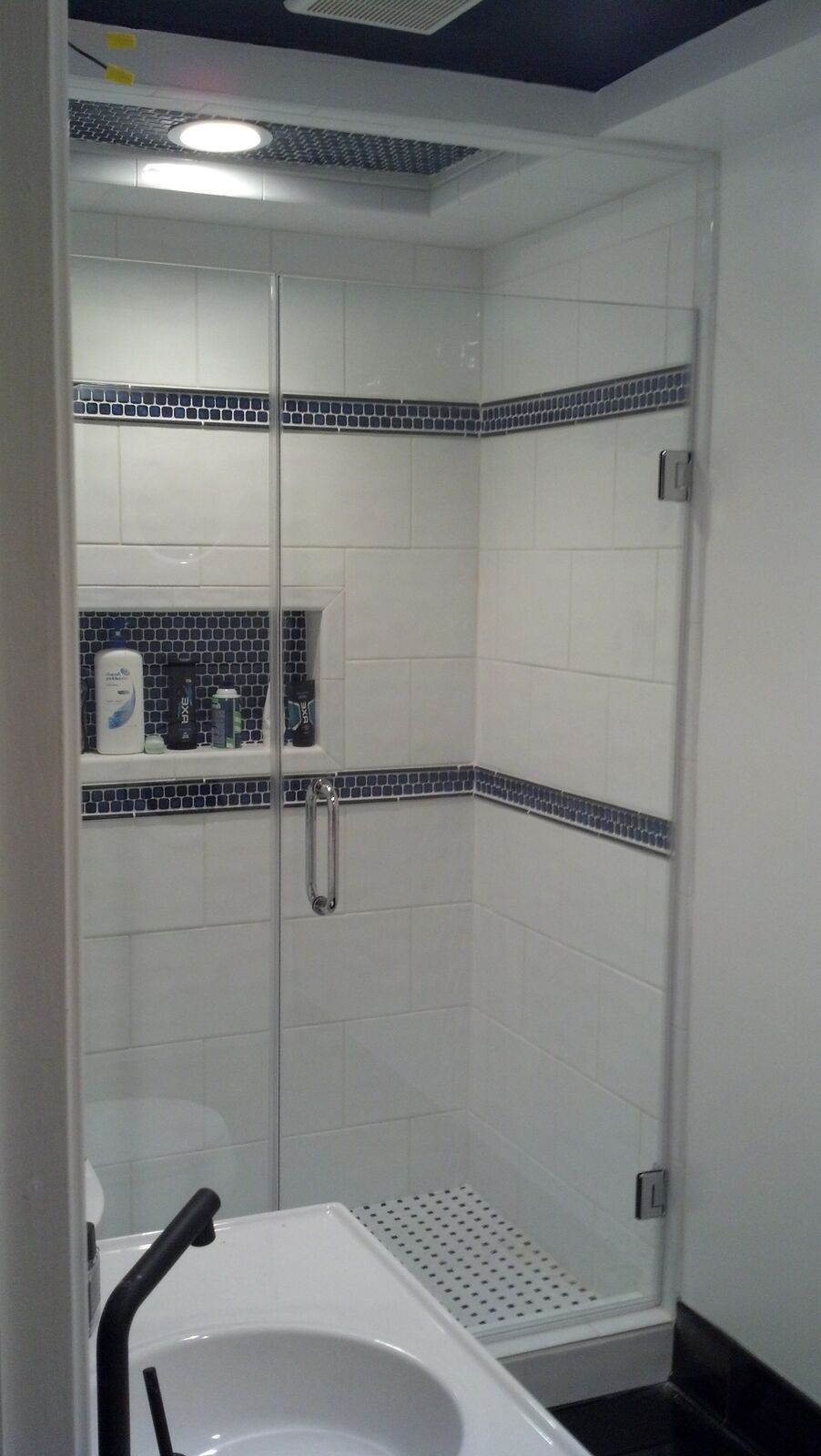 A full-height white-tiled shower with a glass door and blue tile bands, beside a tub
